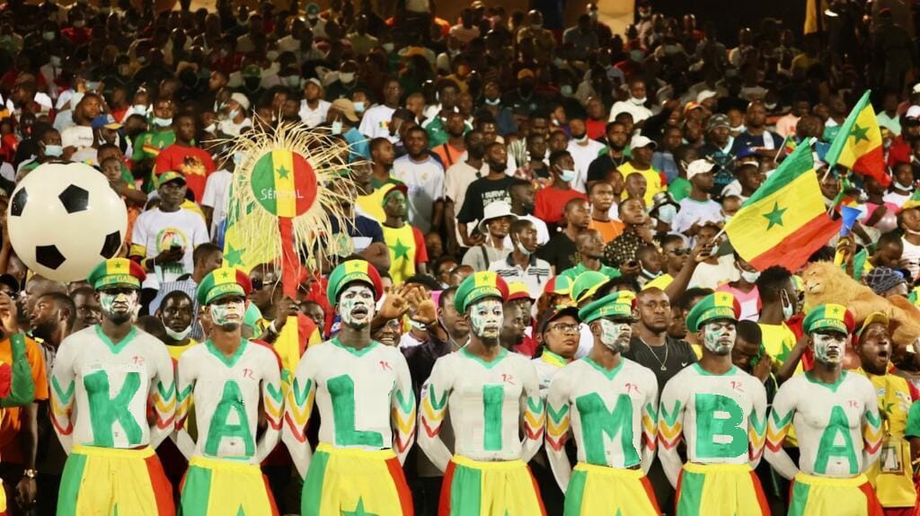 Arquibancada lotada com torcedores da seleção do Senegal durante uma partida de futebol. Em primeiro plano, um grupo de torcedores usa roupas nas cores verde, amarelo e vermelho e forma, com letras pintadas no corpo, a palavra "KALIMBA". Ao fundo, centenas de pessoas seguram bandeiras do Senegal e objetos de torcida, criando uma grande celebração coletiva do futebol africano.